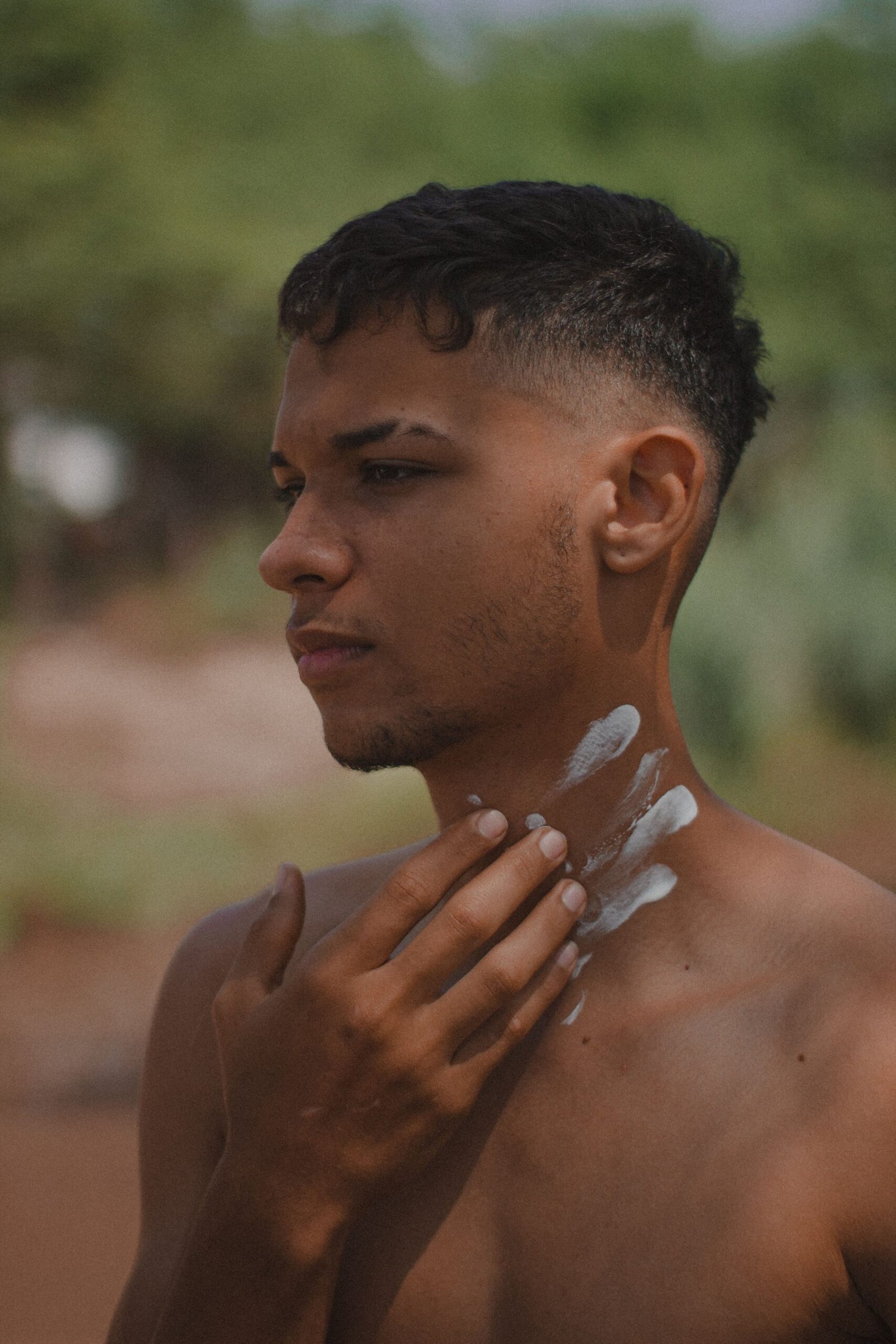 A portrait of a young shirtless man applying skincare on his neck in an outdoor natural setting.